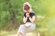 Young woman sitting on a bench praying with Bible in lap