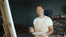 Portrait of a young happy artist in glasses with a brush near the canvas. The man smiles and poses. Creative everyday life.