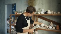 The young potter is doing inventory looking at ceramics and taking notes in workshop. Family business accounting and traditional crafts concept.