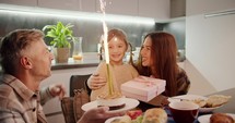 Happy little girl in a cream sweater celebrates her birthday with her parents and blows out a large bright candle on her cake while sitting at the festive table at dinner in the evening in the kitchen