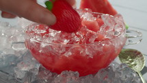 Close up of a transparent glass bowl, standing on pieces of ice, filled with watermelon and ice. Woman's hand placing one strawberry in a bowl with dessert