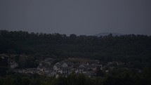 A row of houses with some windows lit against a forested background as night approaches