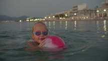 A six-year-old blonde girl, wearing swimming goggles, happily swims in the twilight sea, holding a rubber ball, with the city lights illuminating the background.