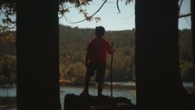 a boy with a walking stick looking out at a lake 