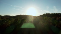 Drone flying high through beautiful mountains and hills covered in green trees with the sunset in the background.