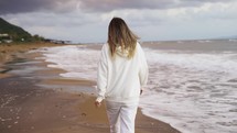 Blonde woman walking along ocean coast alone, rear view.