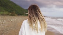 Backview portrait of blonde woman walking along ocean coast, talking by phone.
