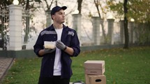 Relaxed courier in blue uniform, enjoying eating food from lunchbox and standing in autumn street near the cardboard boxes.