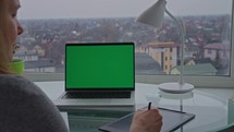 Woman taking notes on a tablet, with a laptop on her desk, overlooking a scenic view