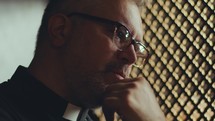 Mature priest sitting inside of confessional, holding hand on chin and having private talk with penitent sitting behind latticed screen. Close-up view
