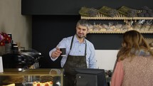 Handsome male barista in black apron giving paper cup takeaway coffe to the female customer. Indoor, bakery.