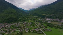 A drone ascends capturing an alpine village embraced by lush mountain slopes, with a serene valley sprawling below and the dramatic sky above