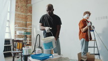 Young African American man pouring paint from bucket into plastic tray as his Caucasian wife preparing roller for house renovation
