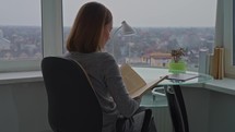 rear view woman sitting on the chair in front of big window reading book at home leisure time for hobby