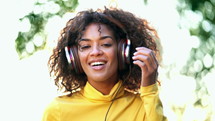 Girl's face in headphones. Young woman in yellow enjoys music while sitting in park.