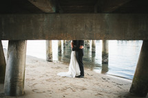 legs and torso of a bride and groom standing under a pier on a beach