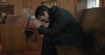 Young, emotional, anxious, and stressed man with long hair and black suit sitting in old church in worship and praying.