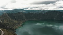 Cinematic drone backwards shot of Quilotoa Crater Lake during cloudy day - Beautiful hiking trails on mountaintop