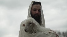 Jesus Christ, the good shepherd of Psalm 23, holding a baby lamb or sheep in his arms while wearing a white, tattered tunic and hood outside in front of cloudy sky during sunset.