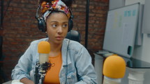 Young African-American woman wearing headphones listening to senior man speaking in mic in the recording studio while hosting audio podcast together. PAN shot