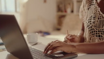 Hands of black girl typing on laptop and making notes at desk in sunlit room, working or studying online from home. Close-up shot
