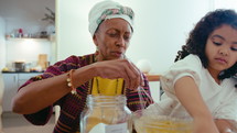 Little black girl adding flour to glass bowl as senior grandmother whisking ingredients and telling recipe, cooking food together in the kitchen at home
