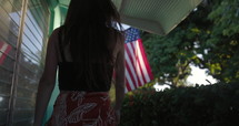 Young woman walks home and touches American flag before going in - from behind