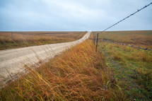Long empty dirt road and barbed wire fence through grassy farmland fields in Kansas during fall