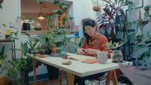 Woman cutting dried leaves with scissors and sticking them to scrapbook pages while making her own herbarium at home office decorated with green plants
