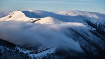 Low inversion clouds drift over snow-capped mountain peaks and hills in the valley below, showcasing the beautiful morning scenery in the national park, with rocks on the mountaintop in the foreground