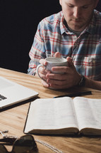 Man holding a coffee cup in prayer over an open Bible on a wooden table.