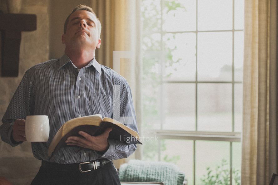 Standing man praying with a Bible and a cup of coffee.