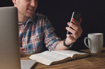 Smiling man holding a cell phone while sitting at a wooden table with an open Bible, coffee cup, and a laptop computer.
