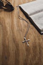 A cross on a chain, sunglasses and an open Bible on a wooden tabletop.