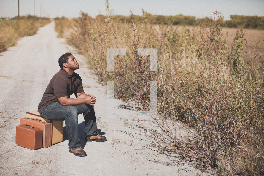man sitting on luggage on a dirt road praying 