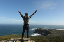woman standing on a mountain top over the ocean with arms raised in praise and worship