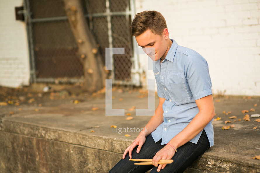 Teen boy sitting on  the sidewalk ledge holding drum sticks.