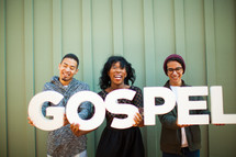 Three smiling people holding a sign saying, "gospel."