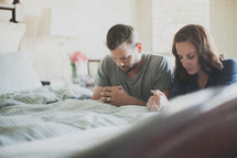 husband and wife praying together at the edge of a bed 