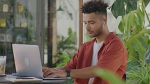 Medium shot of young male freelancer in wireless earphones working on laptop at table in outdoor coffeeshop with notebook, pen and glass of latte on it
