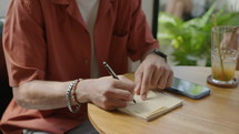 Tilt up shot of young businessman in casual outfit and glasses sitting at cafe table with cocktail and smartphone and taking notes in copybook
