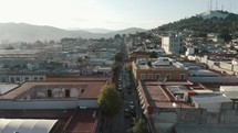 Several cars wait at a crosswalk as a person quietly crosses one of Oaxaca's long linear streets between residential blocks during golden hour in Mexico. Backwards drone dolly shot