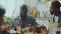 Senior African American man telling a story to family members as they eating and drinking on holiday dinner at home
