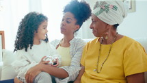 Cheerful African-American woman sitting with little daughter and senior grandmother on sofa in sunlit living room, smiling and chatting when having family time at home
