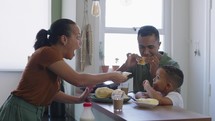 Cheerful family making breakfast while husband and little son eating and drinking juice at kitchen table