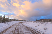 Vehicle tracks in snow on a dirt road near mountains and trees during winter with colorful clouds at sunset 