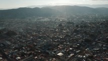 Large square blocks of housing between the huge long linear streets of the Mexican city of Oaxaca with the high green mountain range in the background surmounted by a bright sun. Wide drone tilt shot