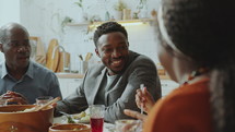 Happy African American family having meal at dinner table, smiling and chatting while celebrating holiday together at home
