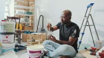 Young African American man sitting on the floor in living room with home improvement supplies and mixing paint colors in cup before painting walls
