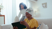 Senior black grandmother sitting on sofa, showing something on digital tablet and talking to excited little girl smiling and jumping beside, spending day together at home
